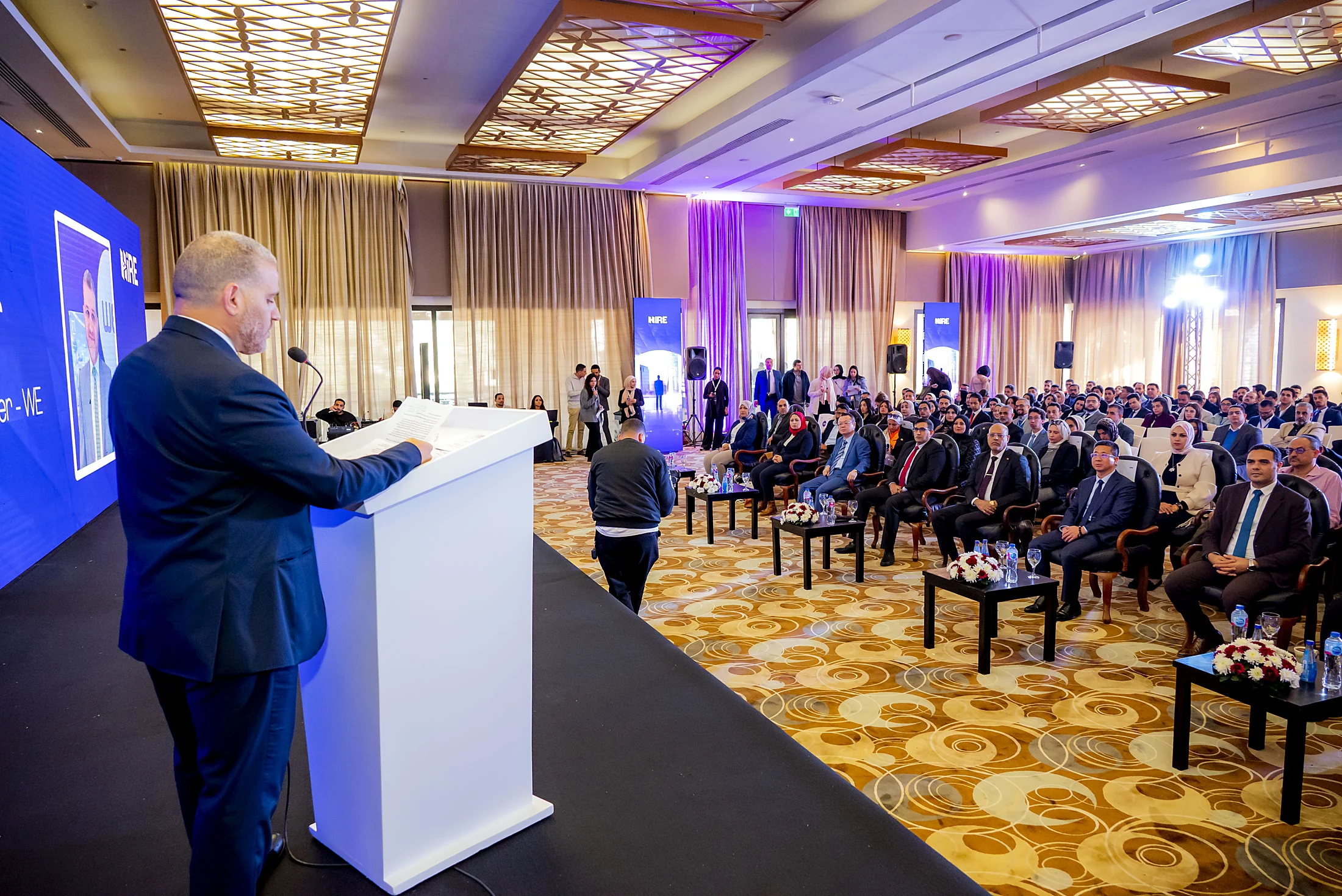 A speaker at the podium addressing a full seated house under the blue HiRE 5 LED