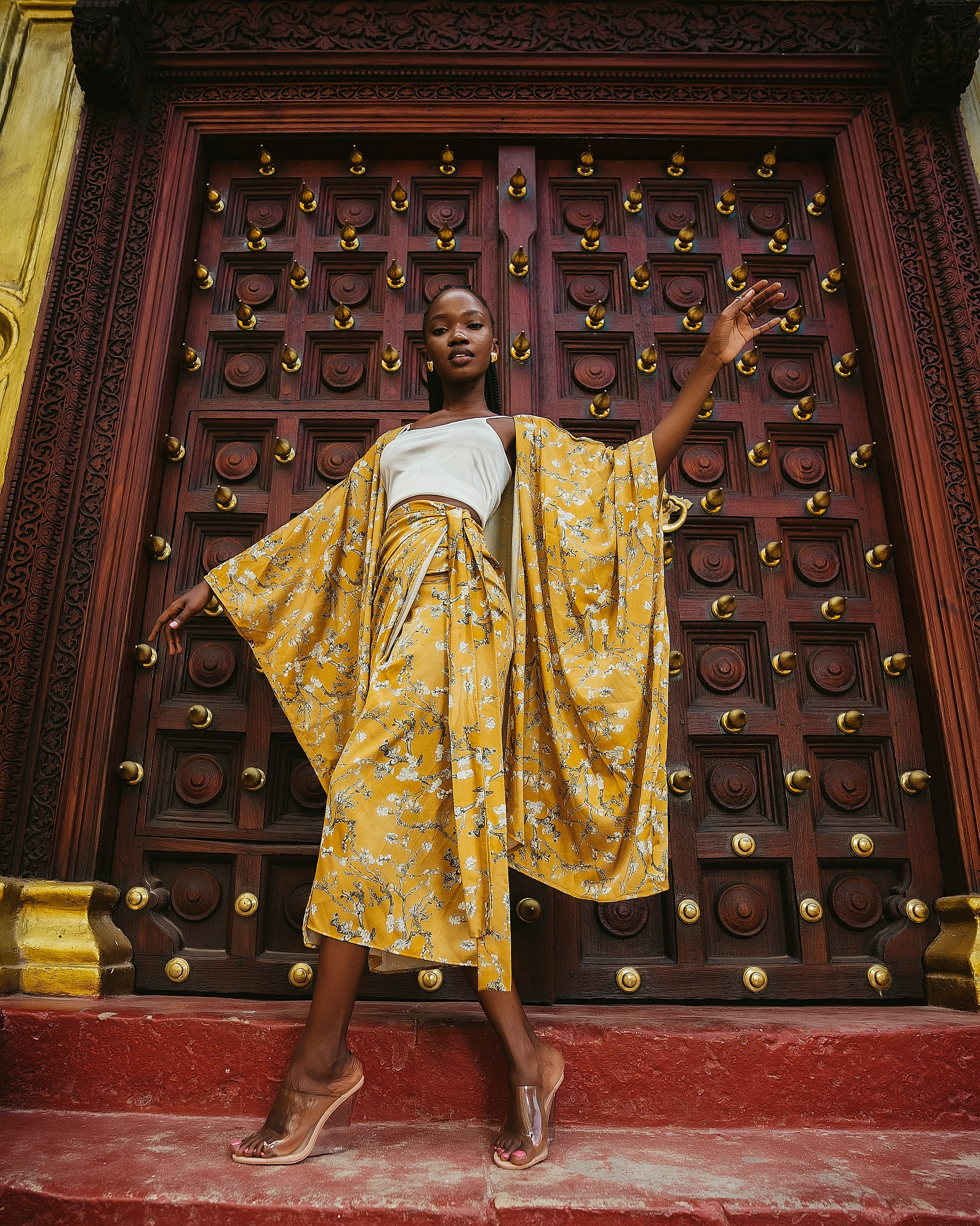 Khokha — a gold botanical robe, arms wide against a carved red Stone Town door, Zanzibar